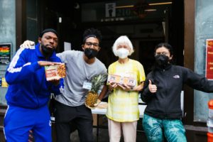 Four masked people smiling at the camera, holding a pre-made meal, a fresh pineapple and eggs - all available at the food pantry.